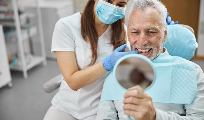 Older man in a dental chair holding up a hand mirror and looking at his new dental implants at Cambridgeside Dental Associates in Cambridge, MA